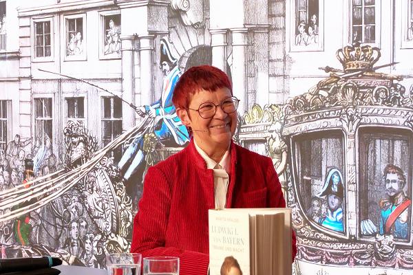 Marita Krauss holding her book “Ludwig I of Bavaria – Dreams and Power” in front of detailed historical illustration at event