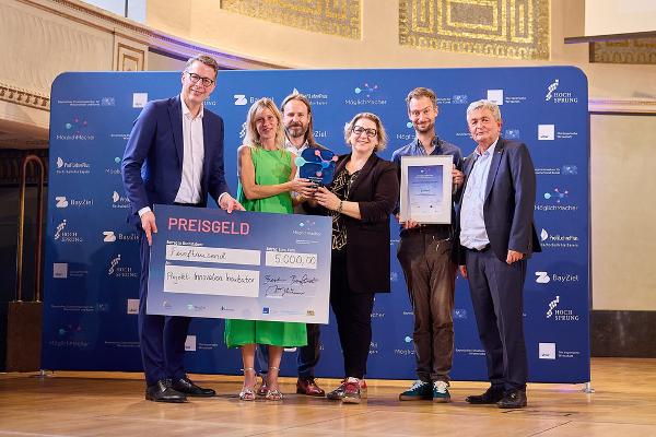 Sechs Personen bei einer Preisverleihung in Bayern präsentieren einen symbolischen Scheck über 5.000 Euro für das Projekt „Innovation Incubator“ sowie eine Auszeichnung vor einer Sponsorenwand.