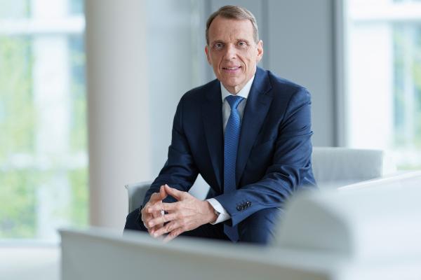 **Alt-Text (Deutsch):** Geschäftsmann im dunkelblauen Anzug mit blauer Krawatte sitzt in modernem, hellem Büro.