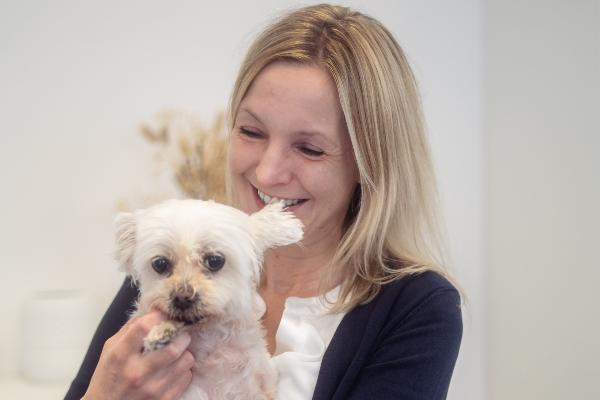 Frau mit blondem Haar hält einen kleinen weißen Malteser-Hund liebevoll im Arm und lächelt.