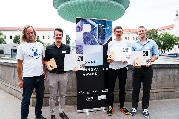 Four winners of the CeNS Nano Innovation Award pose with their certificates and wooden trophies in front of the event banner at the fountain on Geschwister-Scholl-Platz in front of the main building of Ludwig Maximilian University (LMU).