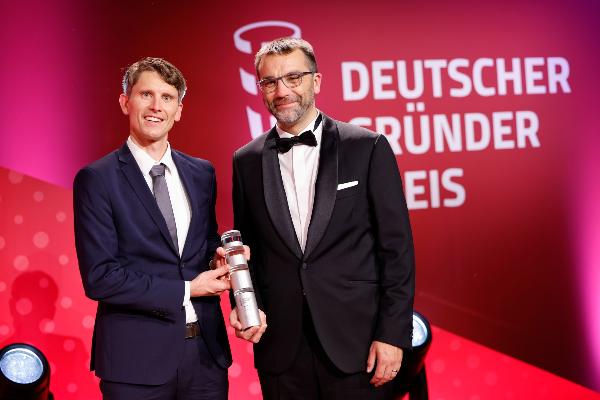 Zwei Männer im Anzug halten den Deutschen Gründerpreis vor rotem Hintergrund mit Logo.