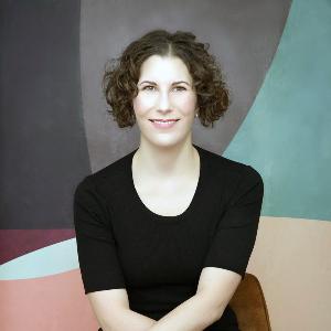 Frau mit kurzen Locken in schwarzem Outfit sitzt auf braunem Stuhl vor farbig-abstraktem Hintergrund.