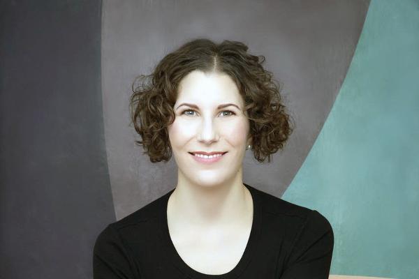 Frau mit kurzen Locken in schwarzem Outfit sitzt auf braunem Stuhl vor farbig-abstraktem Hintergrund.