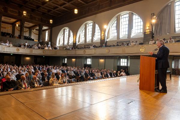 Ein Redner hält eine Ansprache vor einem vollen Saal mit zweistöckigem Publikum in einem eindrucksvollen historischen Gebäude.