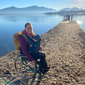 Anna Schaffelhuber in wheelchair with baby carrier enjoying sunny lakeside view with mountain backdrop. | © Privat Anna Schaffelhuber in wheelchair with baby carrier enjoying sunny lakeside view with mountain backdrop.