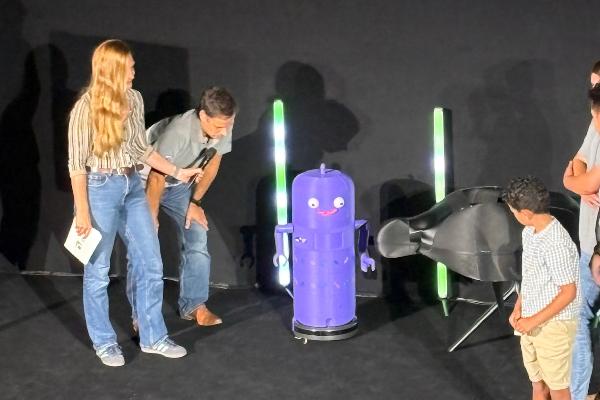 A child, two presenters, and a purple robot stand together on stage.