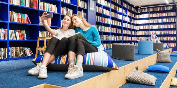 Zwei junge Frauen sitzen in einer Bibliotthek und nehmen ein Selfie auf | © picture alliance / Westend61 | William Perugini Zwei junge Frauen sitzen in einer Bibliotthek und nehmen ein Selfie auf