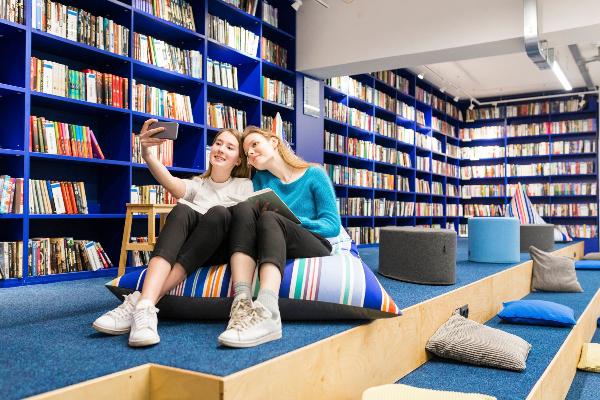 Two young women are sitting in a library library taking a selfie