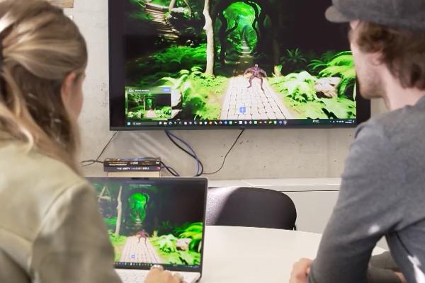 Zwei Personen betrachten 3D-Spielszene auf Laptop und großem Monitor, interaktive Gaming-Umgebung mit grünem Waldpfad.