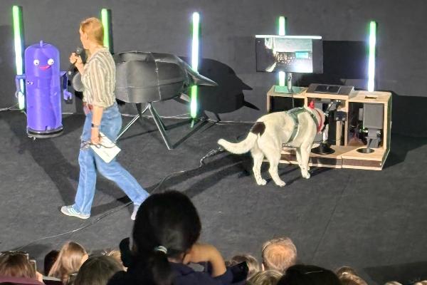 A dog investigates a robot on stage while a presenter introduces the project to an audience.