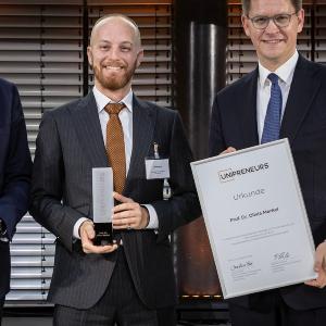 Drei Männer in Anzügen stehen nebeneinander und halten Urkunden und eine Trophäe mit der Aufschrift ´UNIPRENEURS´. Der Mann in der Mitte hält eine gläserne Trophäe, während der Mann rechts eine Urkunde mit dem Namen Prof. Dr. Olivia Merkel präsentiert. Im Hintergrund sind moderne Metalllamellen zu sehen.