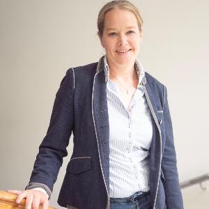 Eine Frau in Jeans, gestreifter Bluse und Blazer steht entspannt an einem Treppengeländer und lächelt in die Kamera.