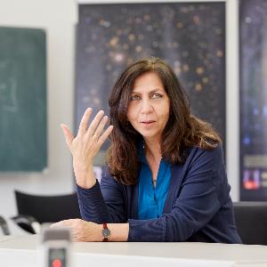 Dr. Cecilia Scorza, gestikulierend im Interview. Im Hintergrund Aufnahmen von Galaxien aus dem Wendelstein-Observatorium