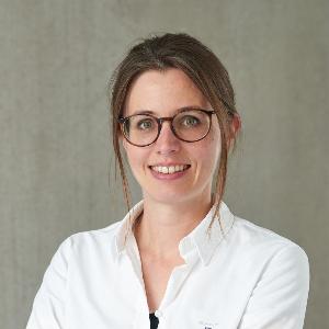 A young woman with brown hair and glasses wears a white shirt and smiles confidently in front of a gray wall.