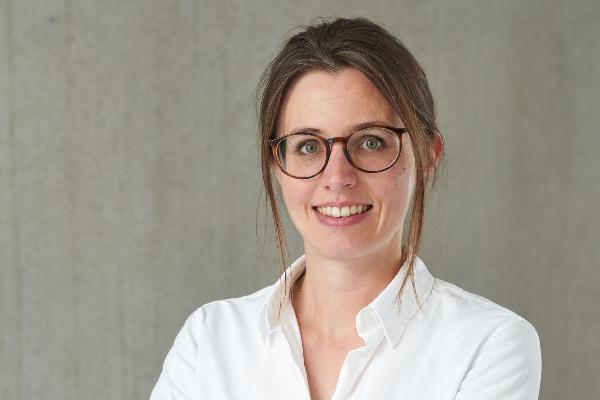 A young woman with brown hair and glasses wears a white shirt and smiles confidently in front of a gray wall.