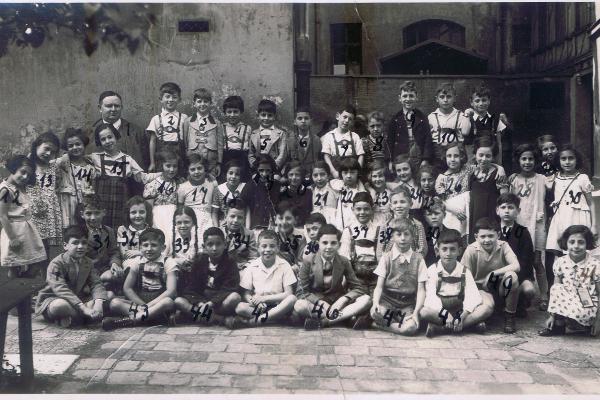 Das Klassenfoto zeigt die 48 Kinder und ihren Lehrer vor der Jüdischen Volksschule in der Herzog-Rudolf-Straße. Die Kinder sind mit Nummer beschriftet, um sie mit der Namensliste auf der Rückseite des Bildes zu verbinden.