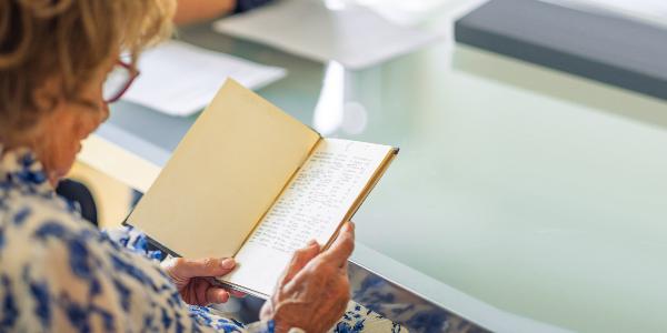 Ältere Frau liest in einem aufgeschlagenen Buch mit handgeschriebenem Text am Tisch.