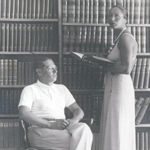 Lion Feuchtwanger, seated, and Martha Feuchtwanger, standing, in front of a densely filled bookshelf. Lion looks somewhat absently in the direction of an open window, Martha looks into the camera and holds an open book in her hands.