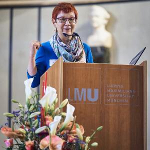 Professor Marita Krauss is giving a lecture at LMU Munich podium, microphones and floral arrangement in the foreground.