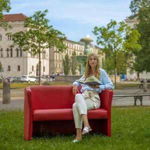 Dr. Marta Diepenrboek auf einer roten Couch vor dem Hauptgebäude der LMU