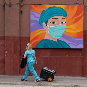 Eine Pflegekraft geht an einem Straßengemälde vorbei, dass eine Pflegekraft mit Mundschutz zeigt. | © IMAGO / ZUMA Wire / Matias Basualdox Eine Pflegekraft geht an einem Straßengemälde vorbei, dass eine Pflegekraft mit Mundschutz zeigt.