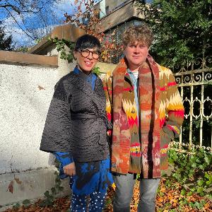 Małgorzata Mirga-Tas und Wojciech Szymański stehen im herbstlichen Garten des Käte Hamburger Kollegs global dis:connect vor einer Mauer und lächeln in die Kamera