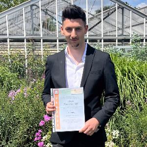 Moritz Gschlößl im schwarzen Anzug hält Urkunde vor Gewächshaus, steht im Garten mit Blumen bei sonnigem Wetter