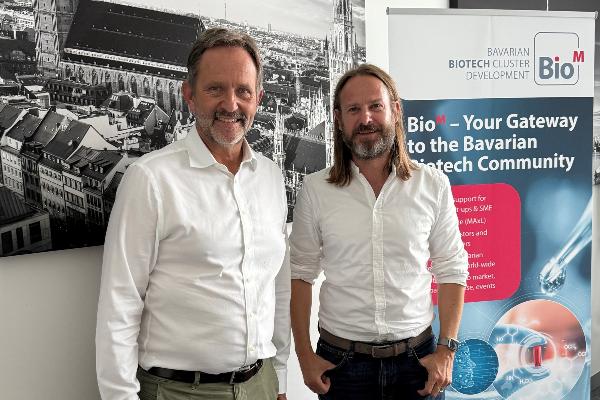 Professor Ralf Huss und Dr. Philipp Baaske in weißen Hemden stehen neben BioM-Banner des Bavarian Biotech Cluster Development vor Münchner Stadtpanorama.