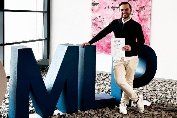 Karim el Marouk hält Zertifikat neben großem PML-Logo in moderner Bürolobby mit Glasfront und pinkem Kunstwerk