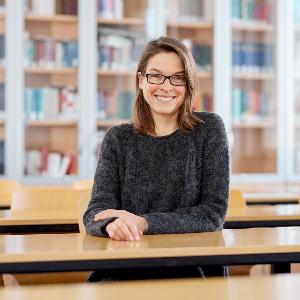 Prof. Barbara Sonnenhauser sitzt an einem Tisch in einer Bibliothek vor einer Bücherwand