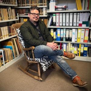 Prof. Dr. Manuel Trummer sitzt auf einem Stuhl vor einer Bücherwand