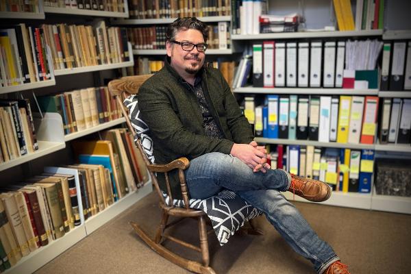 Prof. Dr. Manuel Trummer sitzt auf einem Stuhl vor einer Bücherwand