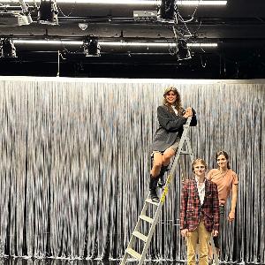 Drei Personen sind auf einer Theaterbühne um eine Leiter gruppiert