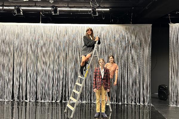 Three people are grouped around a ladder on a theater stage
