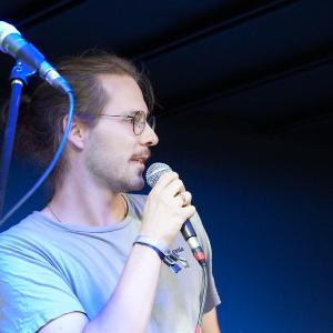 Auf dem Bild ist Philip-Johann Moser zu sehen, der in ein Mikrofon spricht, das er in der Hand hält. Er blickt nachts rechts, trägt eine Brille-