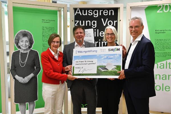 Four people hold a symbolic cheque for €143,000 for the LMU project “Preserve Memory – Shape the Future” in front of information panels.