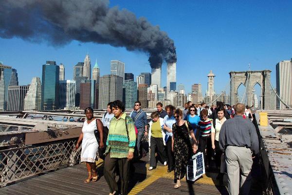 On September 11, 2001, pedestrians cross the Brooklyn Bridge in front of the burning twin towers of the World Trade Center in New York.