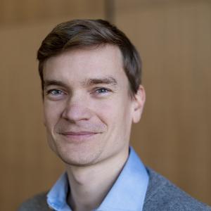 Porträtfoto von Brendan Röder im grauen Pullover und blauen Hemd, der freundlich in die Kamera lächelt, vor einem hölzernen Hintergrund.