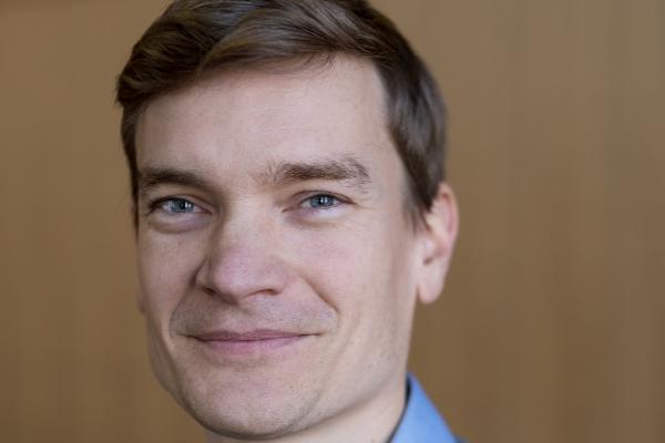 Porträtfoto von Brendan Röder im grauen Pullover und blauen Hemd, der freundlich in die Kamera lächelt, vor einem hölzernen Hintergrund.