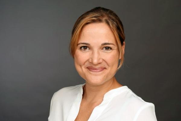 Porträt einer lächelnden Frau mit blonden Haaren im Dutt, in weißer Bluse vor grauem Hintergrund.