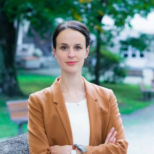Junge Frau mit braunem Blazer steht selbstbewusst mit verschränkten Armen im Freien vor einem Parkweg. | © Tom J.M. Hauzenberger Junge Frau mit braunem Blazer steht selbstbewusst mit verschränkten Armen im Freien vor einem Parkweg.