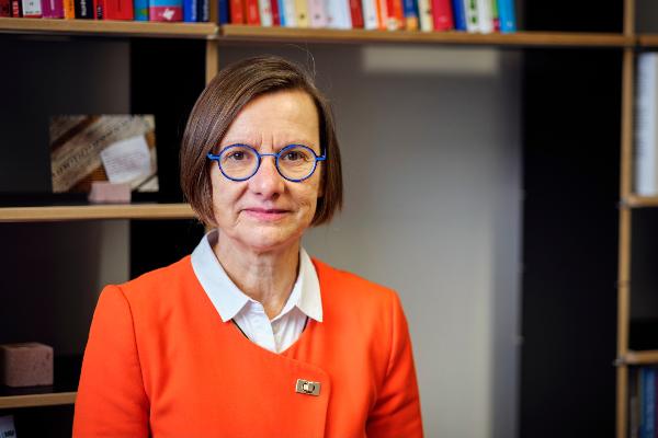 Professor Miriam Schambeck in front of a bookshelf. She is wearing glasses and an orange costume.