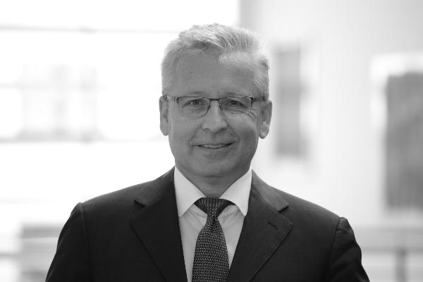 Black and white portrait of Professor Keppler wearing glasses and a suit, standing in front of a bright, modern office background