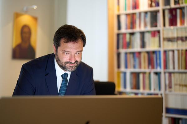 Professor Stefanos Athanasiou blickt konzentriert auf einen Monitor, halbrechts im Hintergrund sieht man eine Ikone mit einer leichten Unschärfe sowie ganz rechts ein volles, sehr aufgeräumtes Bücherregal.