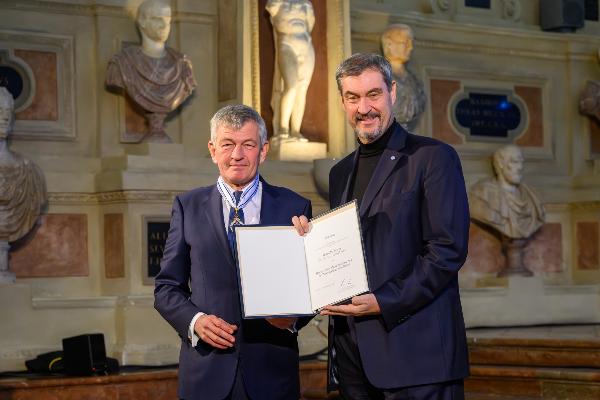 Professor Bernd Huber und Dr. Ministerpräsident Markus Söder in festlichem Saal präsentieren eine Auszeichnung vor historischen Statuen für eine offizielle Ehrung. Bernd Huber hält die Auszeichnungsurkunde und den Maximiliansorden.