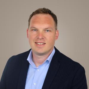 Porträt von Professor Paradies in hellblauem Hemd und dunklem Blazer vor beigem Hintergrund, professionelles Business-Foto.