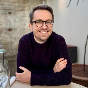Professor Neil Dewar wearing glasses and a dark sweater sitting at a wooden table in a modern, light-filled café or restaurant. The stylish interior with natural daylight, exposed stone wall, and minimalist design creates a welcoming atmosphere for dining, business meetings, and leisure in an urban setting.