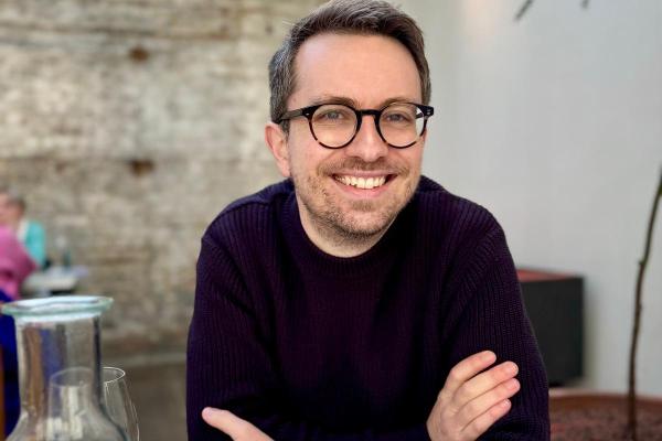 Professor Neil Dewar wearing glasses and a dark sweater sitting at a wooden table in a modern, light-filled café or restaurant. The stylish interior with natural daylight, exposed stone wall, and minimalist design creates a welcoming atmosphere for dining, business meetings, and leisure in an urban setting.