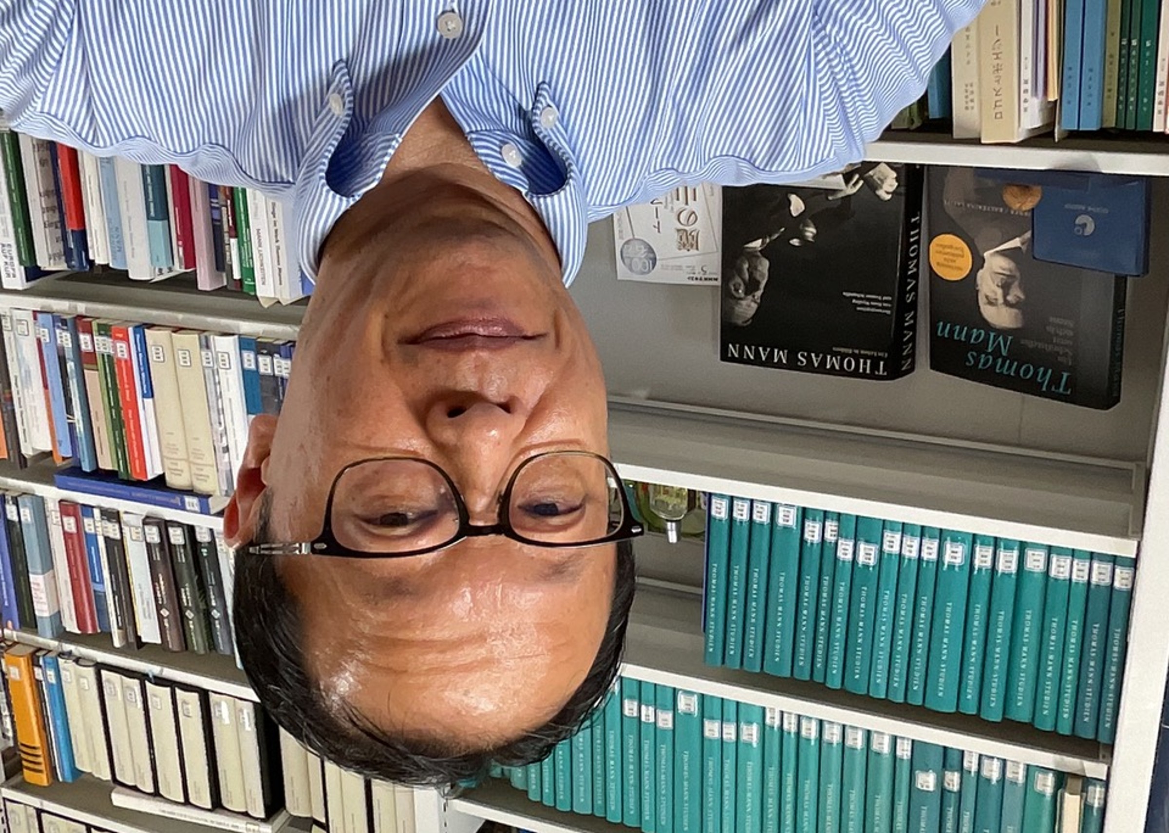 Portrait of Professor Oguro in a blue shirt in front of a bookshelf filled with numerous books in green and white covers. In the foreground, two books by Thomas Mann are visible, one with a photo of him on the cover. The man is wearing glasses and looking into the camera.
